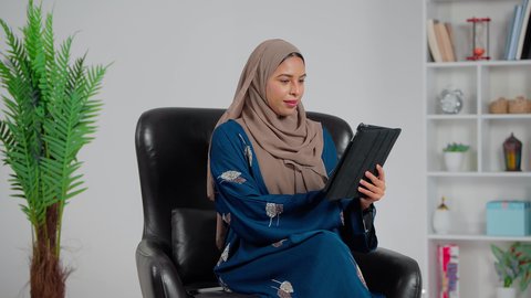 Raising the hand with expressions of greeting and peace, an Arab Gulf Saudi woman wearing an abaya and hijab sits on a chair and uses a tablet to make a video call, accomplishing work tasks remotely, gestures of happiness and joy.