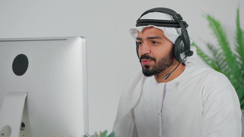 Customer service concept, remote speaking programs, use of headphones and advanced technologies in work, communicating with customers to solve problems and complaints, answering customer questions, close-up of a Saudi Arabian Gulf employee wearing a headset sitting in front of a computer