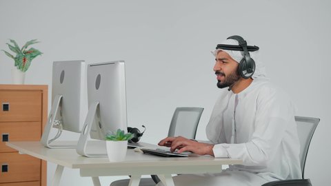 برامج التحدث عن بعد، استخدام الهيدفون والتقنيات المتطورة في العمل، التواصل مع الزبائن لحل المشكلات والشكاوي، الاجابه على اسئلة العملاء، تصوير من الجانب موظف عربي خليجي سعودي يضع سماعة الرأس الصوتية يجلس امام الحاسب الالى يعمل في مجال خدمة العملاء ،النقر على لوحة المفاتيح