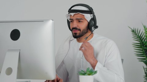 Using headphones and advanced technologies at work, communicating with customers to solve problems and complaints, answering customer questions, an Arab Gulf Saudi employee wearing a headset sitting in front of the computer working in the field of customer service, making a call through the computer