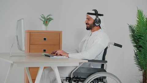 Responding to inquiries and questions, using headphones and advanced technologies at work, a side view of an Emirati Gulf Arab man wearing a kandura and ghutra, wearing headphones, sitting in a wheelchair, working in the customer service department, communicating with customers to solve problems and complaints, clicking on the keyboard