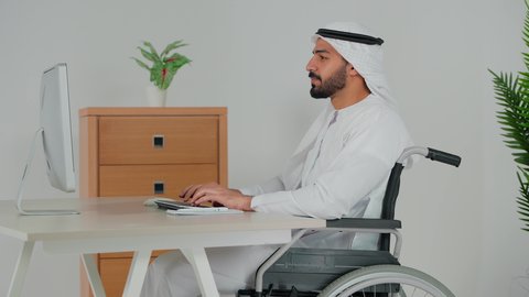 مفهوم إدارة الأعمال، التفاهم والتعاون بين الزملاء لتحقيق أهداف الشركة، إنجاز المهام عبر الحاسب الالى، متابعة إستراتيجية العمل، تصوير من الجانب لرجل عربي خليجي اماراتي يرتدي الكندورة و الغترة يجلس على كرسي متحرك يقوم بامضاء اوراق العمل