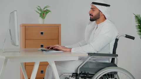 Understanding and cooperation between colleagues to achieve company goals, completing tasks via computer, business management concept, discussing projects and following up on business strategy, side view of an Emirati Gulf Arab man wearing a kandura and ghutra sitting in a wheelchair tapping on a keyboard