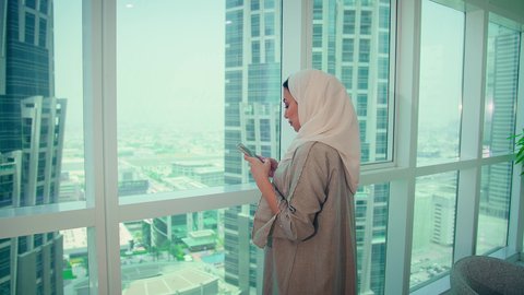 موظفة عربية خليجية اماراتية ترتدي العباءه والحجاب تنظر من خلال الشرفة الهاتف المحمول داخل المكتب، الشركات والمكاتب في الامارات العربية المتحدة، الاطمئنان على الاهل والاصدقاء، استخدام جهاز تقني حديث ومتطور، الدردشة عبر الانترنت