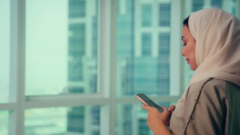 الدردشة عبر الانترنت، الاطمئنان على الاهل والاصدقاء، استخدام جهاز تقني حديث ومتطور، موظفة عربية خليجية اماراتية ترتدي العباءه والحجاب تنظر من خلال الشرفة الهاتف المحمول داخل المكتب، الشركات والمكاتب في الامارات العربية المتحدة