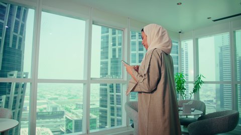 Checking on family and friends, using a modern and advanced technical device, an Emirati Gulf Arab employee wearing an abaya and hijab looking through the balcony at a mobile phone inside the office, companies and offices in the United Arab Emirates, chatting online.