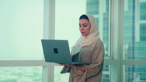 الشركات والمكاتب في الامارات العربية المتحدة،مهنة ووظيفة ادارية مكتبية، استخدام جهاز تقني حديث ومتطور، موظفة عربية خليجية اماراتية ترتدي العباءه والحجاب تنظر من خلال الشرفة تقوم بالعمل على الحاسوب المحمول داخل المكتب،  إدارة شؤون الأعمال وتنظيمها