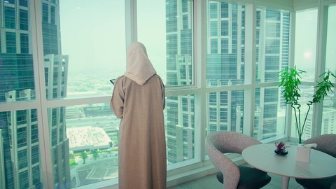 The concept of business management, an Arab Gulf Emirati woman looking through the balcony, studying new projects, economic growth and financial investment, analyzing and auditing project data, writing and taking notes.