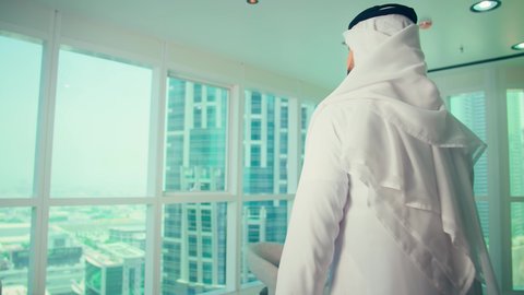 تصوير من الخلف لرجل عربي خليجي إماراتي يرتدي الكندورة والغترة يقف داخل المكتب وينظر من خلال النافذة نحو الأبراج  الحديثة،  بيئة عمل احترافية ، مفهوم التفكير المستقبلي والتخطيط للأهداف، ايماءات الطموح والنجاح