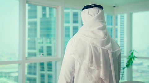 The concept of future thinking and goal planning, a rear view of an Emirati Gulf Arab man wearing a kandura and ghutrah standing inside an office looking out the window at modern towers, a professional work environment, gestures of ambition and success.