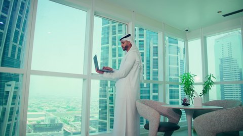 إدارة شؤون الأعمال وتنظيمها، مهنة ووظيفة ادارية مكتبية، استخدام جهاز تقني حديث ومتطور، موظف عربي خليجي اماراتى يرتدي الكندورة والغترة ينظر من خلال الشرفة يقوم بالعمل على الحاسوب المحمول داخل المكتب، الشركات والمكاتب في الامارات العربية المتحدة