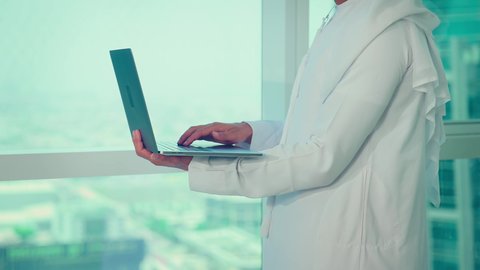 مهنة ووظيفة ادارية مكتبية، استخدام جهاز تقني حديث ومتطور، موظف عربي خليجي اماراتى يرتدي الكندورة والغترة ينظر من خلال الشرفة يقوم بالعمل على الحاسوب المحمول داخل المكتب، الشركات والمكاتب في الامارات العربية المتحدة، إدارة شؤون الأعمال وتنظيمها