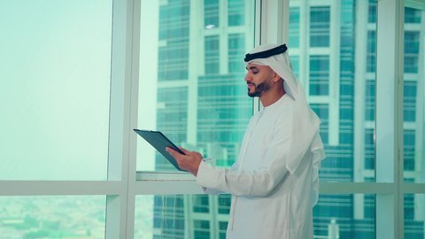 Writing and taking notes, studying new projects, economic growth and financial investment, business management concept, an Emirati Gulf Arab man wearing a kandura and ghutra looking through the balcony, analyzing and checking project data