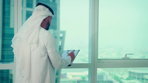 النمو الاقتصادي والاستثمار المالي، دراسة مشاريع جديدة، مفهوم إدارة الأعمال ، رجل عربي خليجي اماراتي يرتدي الكندورة والغترة ينظر من خلال الشرفة ، تحليل وتدقيق بيانات المشروع كتابه وتدوين الملاحظات