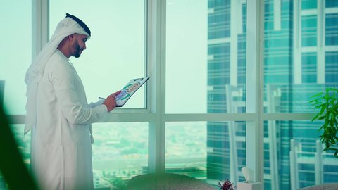 دراسة مشاريع جديدة، النمو الاقتصادي والاستثمار المالي،مفهوم إدارة الأعمال ، رجل عربي خليجي اماراتي يرتدي الكندورة والغترة ينظر من خلال الشرفة ، تحليل وتدقيق بيانات المشروع كتابه وتدوين الملاحظات