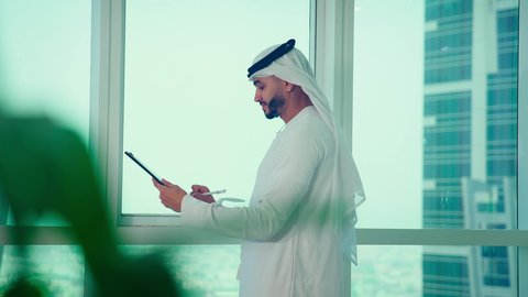 رجل عربي خليجي اماراتي يرتدي الكندورة والغترة ينظر من خلال الشرفة ، ، كتابه وتدوين الملاحظات، دراسة مشاريع جديدة، النمو الاقتصادي والاستثمار المالي،مفهوم إدارة الأعمال ،تحليل وتدقيق بيانات المشروع