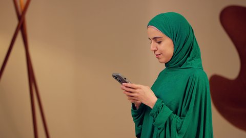 A lantern and decorations for the holy month, an Arab Gulf Emirati woman wearing an abaya and hijab holding a Ramadan lantern and using a mobile phone, taking commemorative selfies, live streaming video, a Saudi woman looking with gestures of joy and happiness, spiritual Ramadan atmosphere.