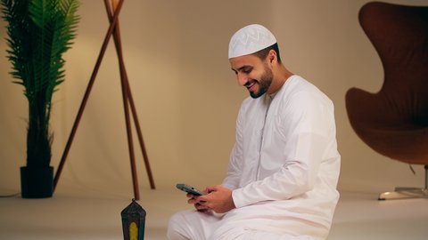 ايماءات السرور و السعادة، اجواء رمضانية روحانية، فانوس وزينة الشهر الكريم ، رجل عربي خليجي اماراتي يرتدي الثوب الابيض عربي يمسك فانوس رمضان ويستخدم الهاتف المحمول، التقاط صور سيلفي تذكارية، فيديو بث مباشر