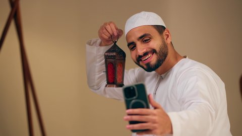 فانوس وزينة الشهر الكريم ، رجل عربي خليجي اماراتي يرتدي الثوب الابيض عربي يمسك فانوس رمضان ويستخدم الهاتف المحمول، التقاط صور سيلفي تذكارية، فيديو بث مباشر، ايماءات السرور و السعادة، اجواء رمضانية روحانية