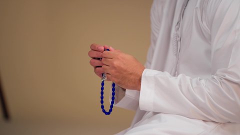 التسبيح والتهليل وذكر الله تعالى، الاجتهاد في العبادات، العبادة والطاعة والتقرب الى الله، تصوير مقرب ليد شاب عربي خليجي اماراتي يرتدي الثوب الأبيض يمسك بيده سبحة من حبات من الخرز الزرقاء