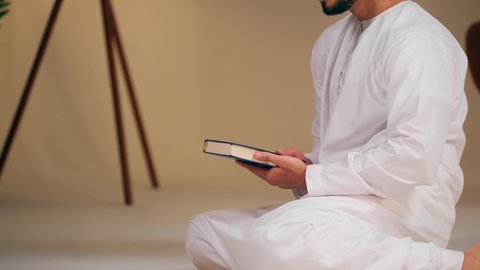 تصوير مقرب لشاب عربي خليجي اماراتي يرتدي الثوب الأبيض والطاقية يحمل بيده القرآن، مفهوم الاجتهاد في العبادة، قراءة كتاب الله وحفظه وتجويده، تلاوة المصحف الشريف، شاب عربي خليجي اماراتي يرتدي الثوب الأبيض والطاقية يحمل بيده القرآن