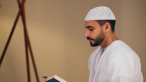 قراءة كتاب الله وحفظه وتجويده، مفهوم الاجتهاد في العبادة، تلاوة المصحف الشريف، شاب عربي خليجي اماراتي يرتدي الثوب الأبيض والطاقية يحمل بيده القرآن