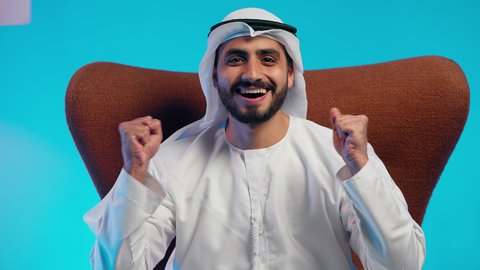 Pride and the feeling of happiness for achieving something, facial gestures with excessive enthusiasm, an Arab Gulf Emirati man wearing a kandura and ghutrah sitting on a comfortable chair raising his hand up for encouragement, expressions of joy and delight.