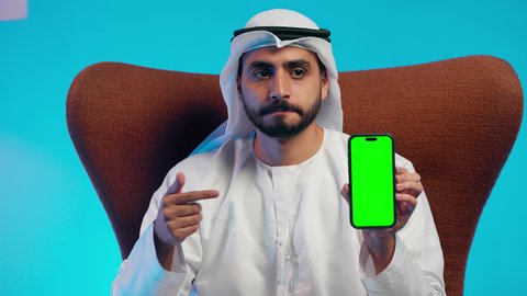 Conducting a live broadcast with family and friends, pointing to a green chroma screen, browsing social media, an Arab Gulf Emirati man wearing a kandura and ghutrah holding a mobile phone in his hand, integrating modern technologies into daily life, using a mobile phone.