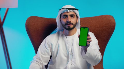 Green chroma screen display, browsing social media, an Arab Gulf Emirati man wearing a kandura and ghutrah holding a mobile phone in his hand, integrating modern technologies into daily life, using a mobile phone, conducting live broadcasts with family and friends, taking commemorative selfie photos.