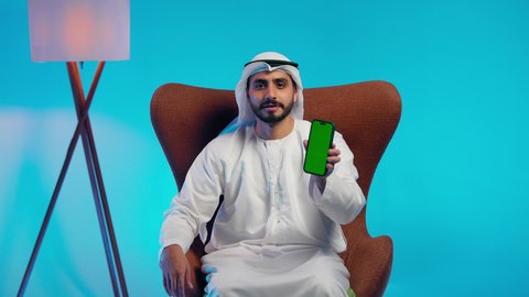Browsing social media, an Arab Gulf Emirati man wearing a kandura and ghutrah holds a mobile phone in his hand, integrating modern technologies into daily life, using a mobile phone, conducting live broadcasts with family and friends, taking commemorative selfie photos.