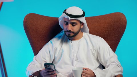 رجل عربي خليجي اماراتي يرتدي الكندورة والغتره يمسك الجوال بيده ، الاستمتاع بالمشروبات الساخنه ، تصفح مواقع التواصل الاجتماعي ، دمج التقنيات الحديثة فى الحياة اليومية ، استخدام الهاتف المحمول ، إجراء بث مباشر مع الأهل والأصدقاء