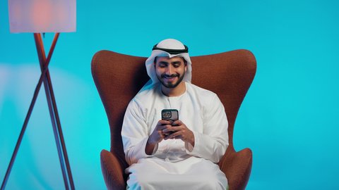 Using a mobile phone, browsing social media, an Arab Gulf Emirati man wearing a kandura and ghutrah holding a phone in his hand, integrating modern technologies into daily life, conducting live broadcasts with family and friends, taking commemorative selfies.