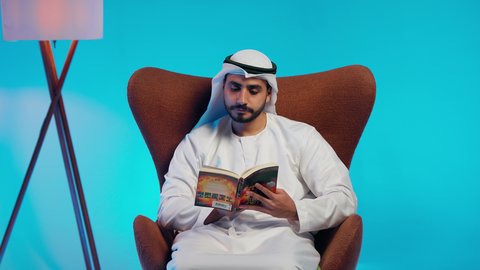 Gestures of attention and focus, spending free time learning and self-education, an Arab Gulf Emirati man wearing a kandura and ghutrah is reading attentively, browsing entertaining illustrated stories, developing reading skills, and being interested in browsing useful books.