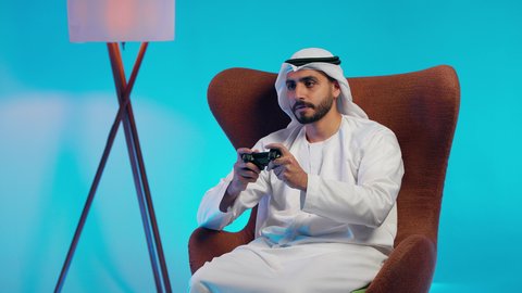 Spending enjoyable and entertaining time, using modern electronic entertainment devices and technologies, interacting and engaging with video games, an Arab Gulf Emirati man wearing a kandura and ghutrah holding a game controller in his hand.
