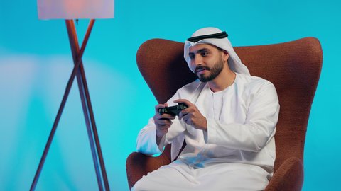 رجل عربي خليجي اماراتي يرتدي الكندورة والغتره يمسك بيده عصا التحكم،التفاعل والاندماج مع ألعاب الفيديو، قضاء وقت ممتع ومسلي، استخدام اجهزة وتقنيات الكترونية ترفيهيه حديثة