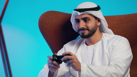 استخدام اجهزة وتقنيات الكترونية ترفيهيه حديثة,  رجل عربي خليجي اماراتي يرتدي الكندورة والغتره يمسك بيده عصا التحكم،التفاعل والاندماج مع ألعاب الفيديو، قضاء وقت ممتع ومسلي