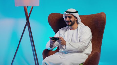 Spending enjoyable and entertaining time, interacting and engaging with video games, using modern electronic entertainment devices and technologies, an Arab Gulf Emirati man wearing a kandura and ghutrah holding a game controller in his hand.