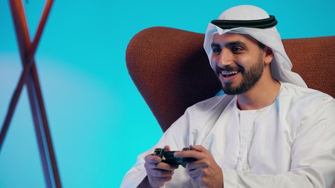 التفاعل والاندماج مع ألعاب الفيديو، قضاء وقت ممتع ومسلي، استخدام اجهزة وتقنيات الكترونية ترفيهيه حديثة,  رجل عربي خليجي اماراتي يرتدي الكندورة والغتره يمسك بيده عصا التحكم