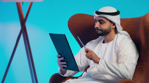 Daily task recording, work-related papers, an Emirati Gulf Arab man wearing a kandura and ghutrah holding white papers and a pen in his hands, a Saudi woman conducting scientific research, reading notes and information, following up on plans and projects.