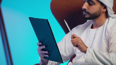 Feasibility study of work, recording daily tasks, an Arab Gulf Emirati man wearing a kandura and ghutrah holding white papers and a pen in his hands, a Saudi woman conducting scientific research, reading notes and information, following up on plans and projects.
