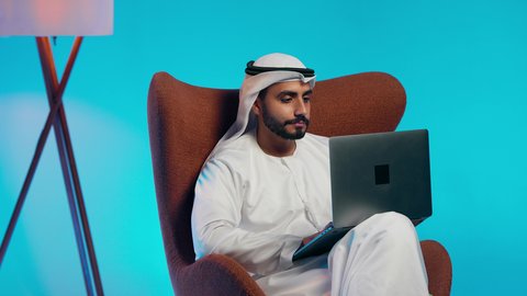 Remote business management, using modern technologies, making video calls with family and friends, an Arab Gulf Emirati man wearing a kandura and ghutrah, with headphones, holding a laptop and looking at it with gestures of happiness.