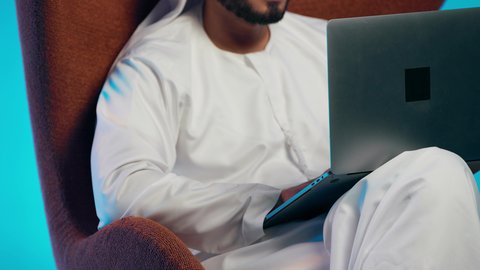 رجل عربي خليجي اماراتي يرتدي الكندورة والغتره وسماعات الرأس يمسك جهاز الحاسوب المحمول وينظر إليه بإيماءات السعادة،استخدام التقنيات الحديثة،إجراء مكالمات الفيديو مع الأهل والأصدقاء، اجتماع عمل عن بعد