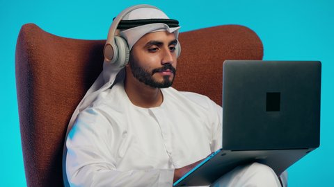 The use of modern technologies, making video calls with family and friends, remote work meetings, an Arab Gulf Emirati man wearing a kandura and ghutrah, with headphones, holding a laptop and looking at it with gestures of happiness.