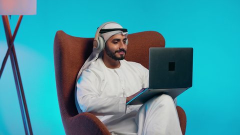 استخدام التقنيات الحديثة ، إجراء مكالمات الفيديو مع الأهل والأصدقاء، اجتماع عمل عن بعد، رجل عربي خليجي اماراتي يرتدي الكندورة والغتره وسماعات الرأس يمسك جهاز الحاسوب المحمول وينظر إليه بإيماءات السعادة