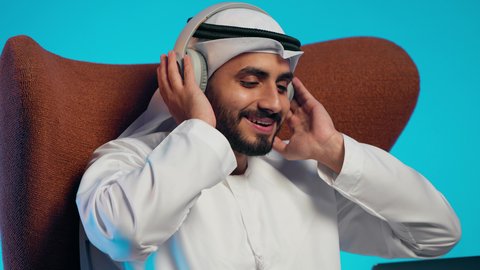Expressions of joy and happiness, using modern technologies, making video calls with family and friends, a remote work meeting, an Emirati Gulf Arab man wearing a kandura and ghutrah, with headphones, holding a laptop and looking at it with gestures of happiness.