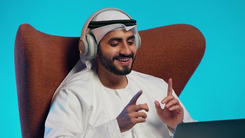إجراء مكالمات الفيديو مع الأهل والأصدقاء، اجتماع عمل عن بعد، رجل عربي خليجي اماراتي يرتدي الكندورة والغتره وسماعات الرأس يمسك جهاز الحاسوب المحمول وينظر إليه بإيماءات السعادة،استخدام التقنيات الحديثة