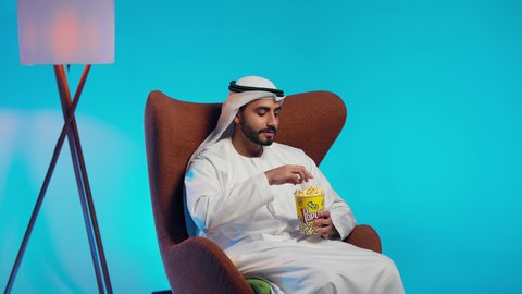 رجل عربي خليجي اماراتي يرتدي الكندورة والغترة يجلس على كرسي مريح ويقوم بتناول الفشار،قضاء وقت الفراغ بمتابعة الافلام و المسلسلات المسلية، تناول سناك صحي و خفيف