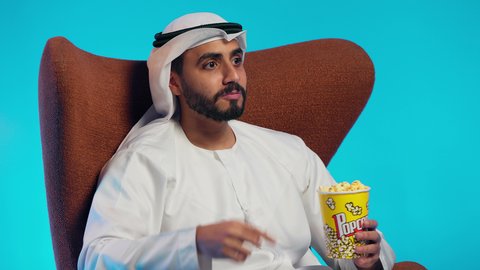 مفهوم التسلية والترفيه، قضاء وقت الفراغ بمتابعة الافلام و المسلسلات المسلية، تناول سناك صحي و خفيف ، رجل عربي خليجي اماراتي يرتدي الكندورة والغترة يجلس على كرسي مريح ويقوم بتناول الفشار
