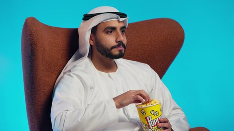 تناول سناك صحي و خفيف ، قضاء وقت الفراغ بمتابعة الافلام و المسلسلات المسلية، رجل عربي خليجي اماراتي يرتدي الكندورة والغترة يجلس على كرسي مريح ويقوم بتناول الفشار