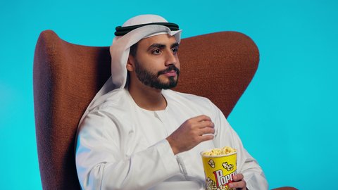 Spending free time watching entertaining movies and series, having a healthy and light snack, an Arab Gulf Emirati man wearing a kandura and ghutrah sits on a comfortable chair and is eating popcorn.